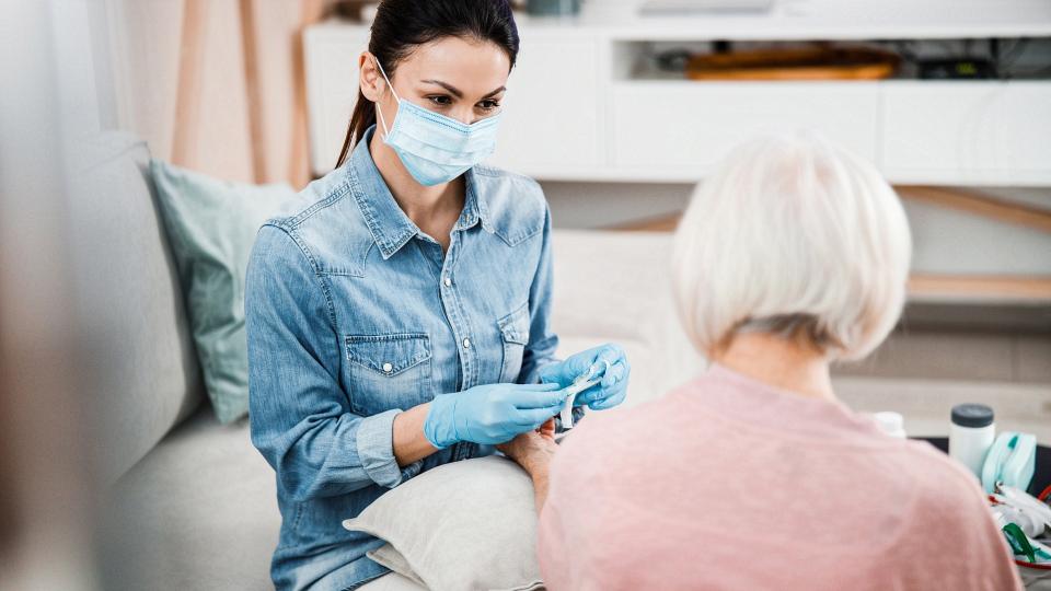 Eine Altenpflegehelferin betreut eine Seniorin im privatem Umfeld. Die Altenpflegehelferin trägt eine Schutzmaske und Gummihandschuhe. Die Seniorin breitet gleichzeitig ihren Arm zur Blutabnahme aus.