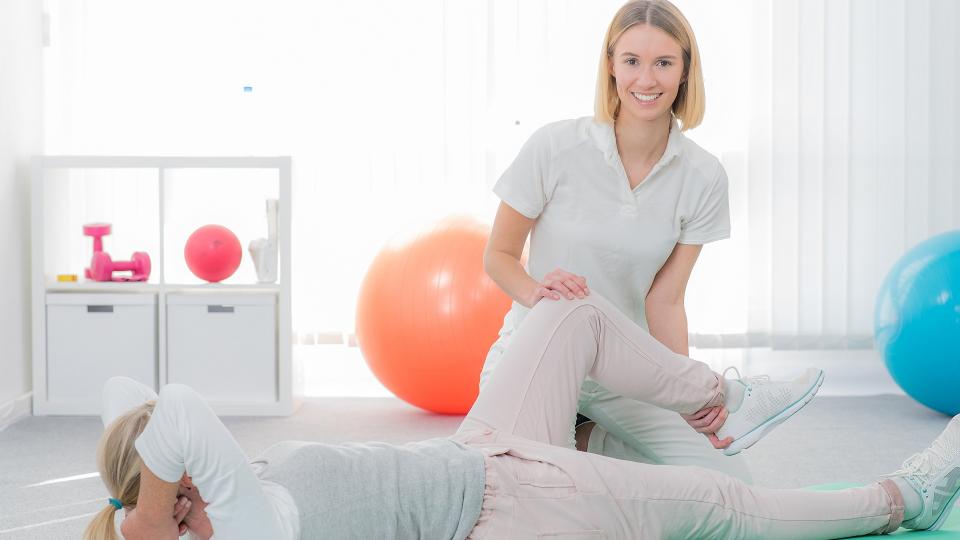 Eine Ergotherapeutin betreut eine Patientin beim Sporttreiben. Die Patientin liegt auf dem Rücken und die Ergotherapeutin beugt das linke Bein der Patientin.  