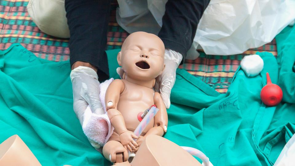 Abgebildet ist eine Übungspuppe in Form eines menschlichen Babys. Gehalten wird die Puppe von einer Person, die transparente Einweghandschuhe trägt. 