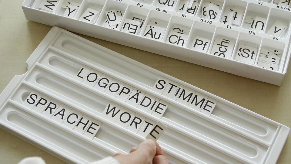 Abgebildet sind zwei weiße Boxen. Die Box im Hintergrund enthält nach dem Alphabet geordnete Buchstabenplättchen. Eine Person hat aus diesen Buchstaben die Worte "Stimme", "Logopädie", "Worte" und "Sprache" in der im Vordergrund befindlichen Box zusammengefügt.