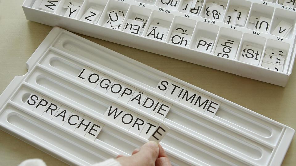 Abgebildet sind zwei weiße Boxen. Die Box im Hintergrund enthält nach dem Alphabet geordnete Buchstabenplättchen. Eine Person hat aus diesen Buchstaben die Worte "Stimme", "Logopädie", "Worte" und "Sprache" in der im Vordergrund befindlichen Box zusammengefügt.  