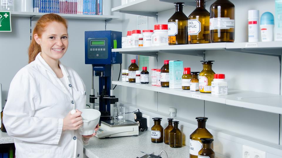 Eine Pharma-Assistentin steht vor einem Regal, auf dem sich toxische Substanzen in mehreren Glasbehältern befinden. Sie hält zudem einen Mörser zum Mischen einiger Substanzen in ihren Händen.