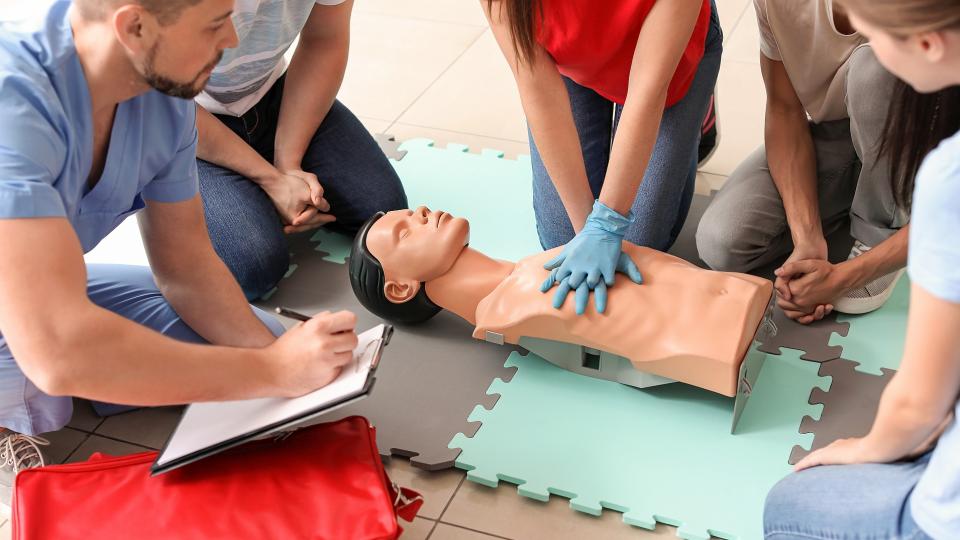 Eine Frau führt an einem menschlichen Dummy eine Herzdruckmassage durch. Ein Mann beurteilt sie dabei. Drei weitere Personen sitzen in einem Halbkreis neben den Protagonisten und schauen konzentriert zu.