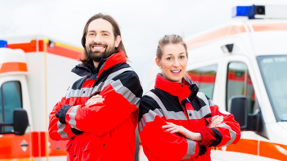 Eine Rettungssanitäterin und ein Rettungssanitäter posieren lächelnd vor zwei Krankenwagen für die Kamera. 