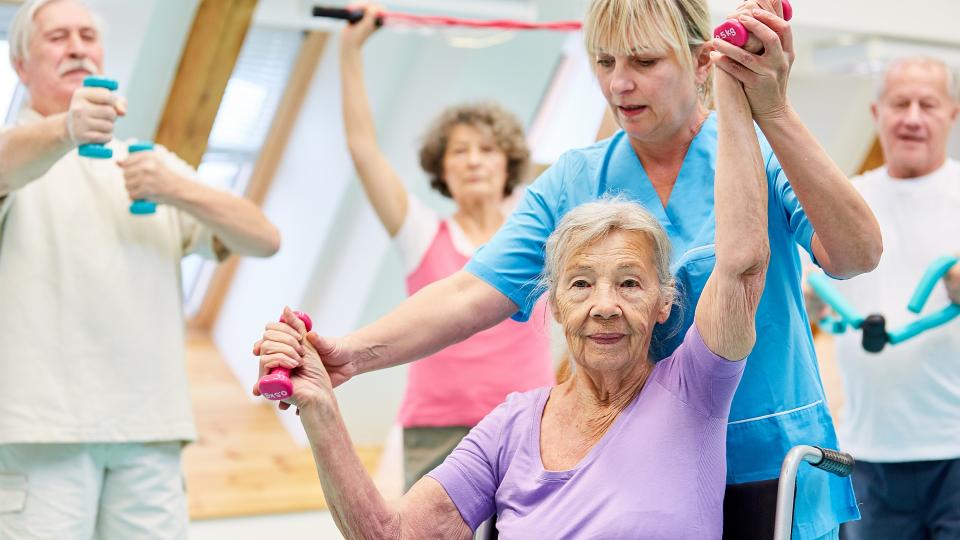 Eine Ergotherapeutin betreut eine ältere Dame beim Sporttreiben. Im Hintergrund befinden sich ältere Personen, die ebenfalls sportliche Aktivitäten ausüben.
