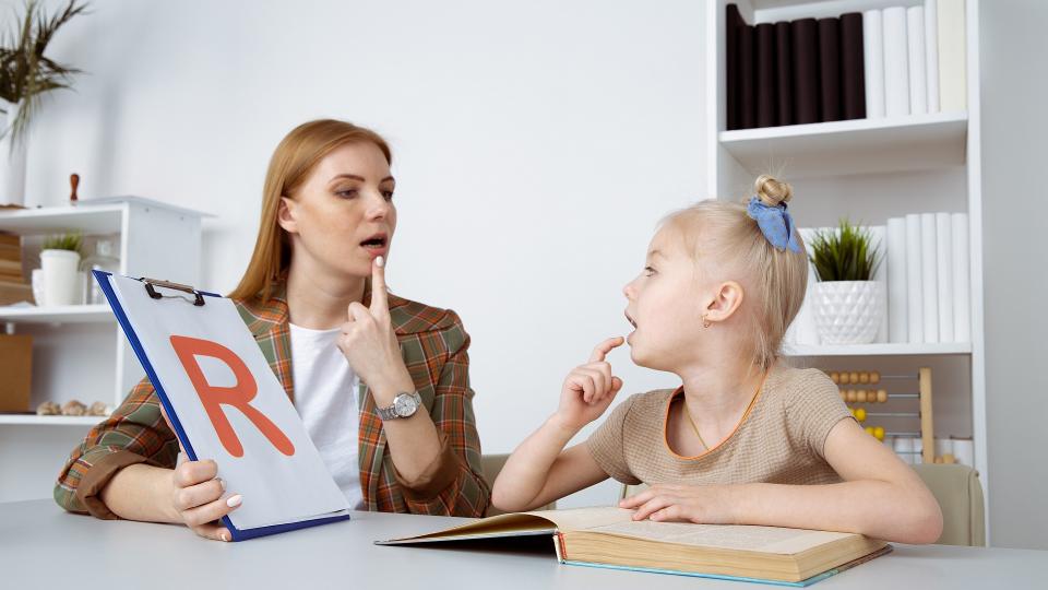 Eine Logopädin übt mit einem Kind die Aussprache des Buchstabens "R". Der Buchstabe steht auf einem Blatt Papier und wird dem Kind deutlich gezeigt. Vor dem Kind befindet sich ein Buch auf einem Schreibtisch. 