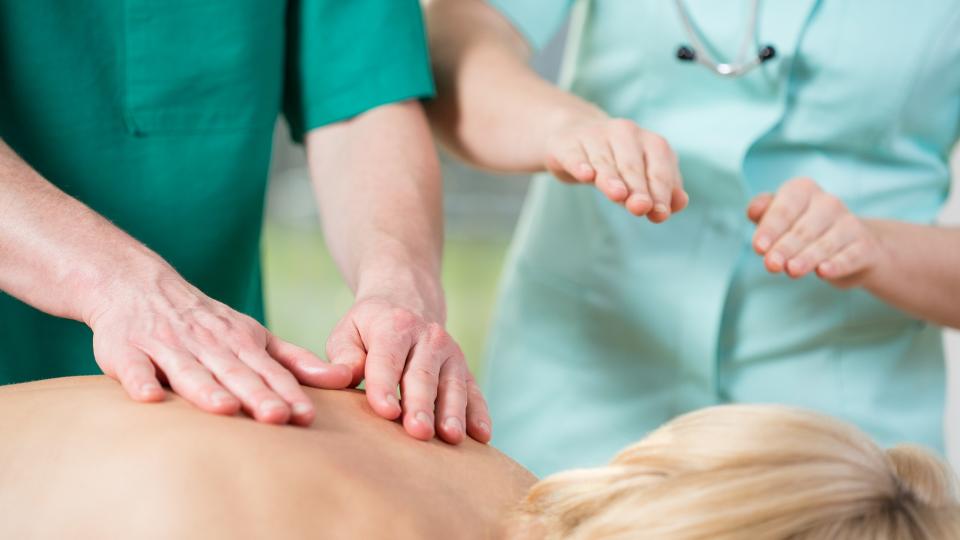 Eine Person massiert den Rücken einer Frau. Neben der massierenden Person steht eine weitere Person, die eine ähnliche Handbewegung andeutet wie die Masseurin beziehungsweise der Masseur ausführt.