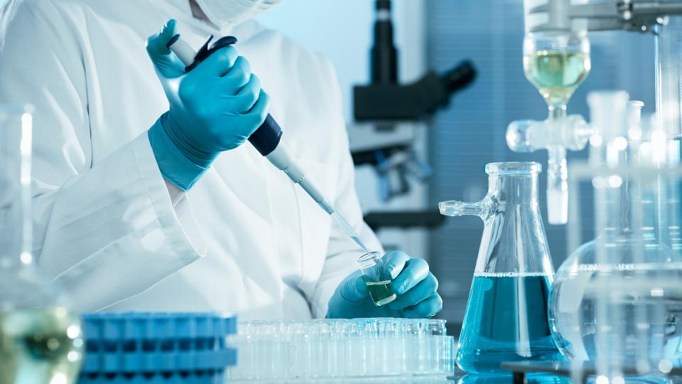 Eine Laboratoriumsassistentin beziehungsweise ein Laboratoriumsassistent füllt mit Hilfe einer Pipette eine Flüssigkeit in ein Reagenzglas. Die Person befindet sich in einem Labor und trägt Schutzkleidung. 