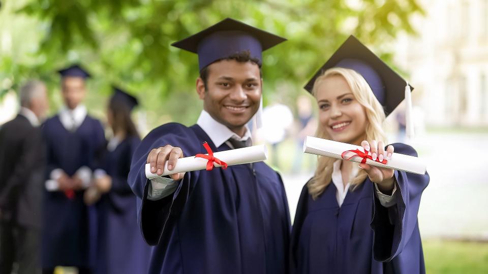 Eine Hochschulabsolventin und ein Hochschulabsolvent zeigen lächelnd und mit ausgestreckten Armen ihre Diplome in Richtung Kamera. Der Mann hat einen Migrationshintergrund.