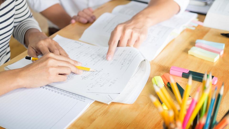 Ein*e Schüler*in und eine Lehrkraft sitzen an einem Schreibtisch, auf dem sich Papier und Büroartikel befinden. Gleichzeitig deuten beide Personen auf ein beschriftetes Blatt Papier.