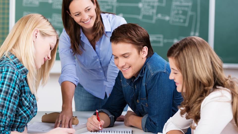 Drei Schülerinnen und Schüler sitzen an einem Schreibtisch in einem Klassenraum und schauen auf Lernunterlagen, die sich in der Mitte des Tisches befinden. Eine Lehrerin steht an diesem Tisch und zeigt mit Ihrer Hand auf die Lernunterlagen.