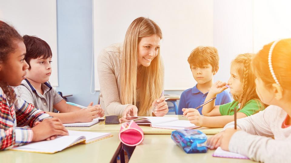 Eine Lehrerin betreut fünf Grundschulkinder in einem Klassenraum. Die Kinder sitzen an drei zusammengestellten Tischen und hören der Lehrerin konzentriert zu. 