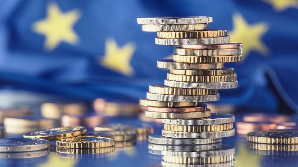 Im Fokus stehen gestapelte Euro-Geldmünzen. Im Hintergrund ist die Europaflagge abgebildet. 