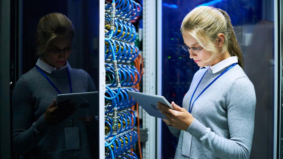 Eine Frau überprüft mit einem Tablet die Funktionalität eines Schaltschranks, aus dem viele Kabel hervorstehen.