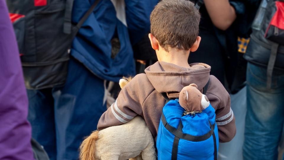 Ein kleiner Junge trägt inmitten vieler Erwachsener einen Rucksack, aus dem ein Kuscheltier zum Vorschein kommt. Ein weiteres Kuscheltier drückt er mit dem linken Arm an seinen Körper. 