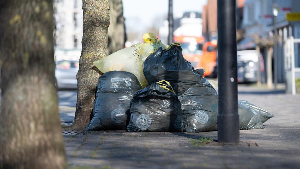 Abgebildet sind mehrere schwarze und gelbe Müllsäcke, die unsortiert am Straßenrand liegen.