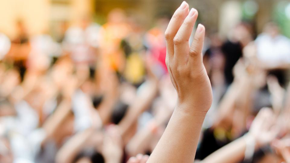 Abgebildet ist ein in die Höhe ausgestreckter Arm. Der Hintergrund ist sehr unscharf. Zu erahnen ist aber, dass die Menschen im Hintergrund ebenfalls die Arme heben.