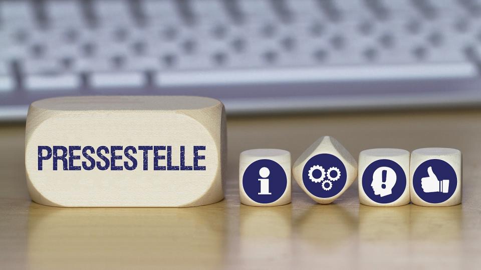 Links abgebildet ist ein kleines Rechteck aus Holz, auf dem "Pressestelle" steht. Rechts davon befinden sich vier kleinere Holzwürfel. Auf einem Würfel ist ein "i" für Information abgebildet, auf den anderen Zahnräder, das Profil eines menschlichen Kopfes mit einem eingearbeiteten Ausrufezeichen und eine Hand mit einem nach oben gerichteten Daumen. Im Hintergrund befindet sich eine Tastatur.