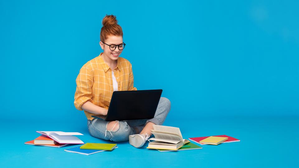 Rothaarige Stundentin sitzt mit einem Laptop auf dem Schoß inmitten von Büchern.