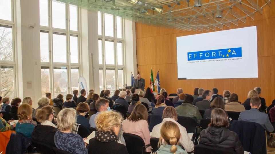 Herr Regierungspräsident Hans-Josef Vogel hält im großen Sitzungssaal der Bezirksregierung Arnsberg einen Vortrag über EFFORT-A. Der Sitzungssaal ist komplett mit Zuhörerinnen und Zuhörern gefüllt.