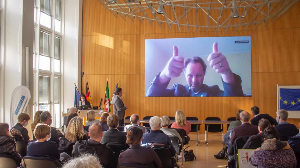 Ein Mann ist live dem Vortrag auf einem großen Bildschirm zugeschaltet. Er zeigt beide Daumen nach oben.