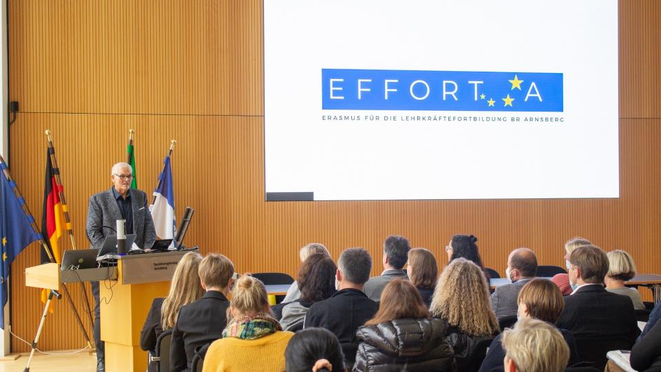 Herr Regierungspräsident Hans-Josef Vogel hält im großen Sitzungssaal der Bezirksregierung Arnsberg einen Vortrag über EFFORT-A. Der Sitzungssaal ist komplett mit Zuhörerinnen und Zuhörern gefüllt.