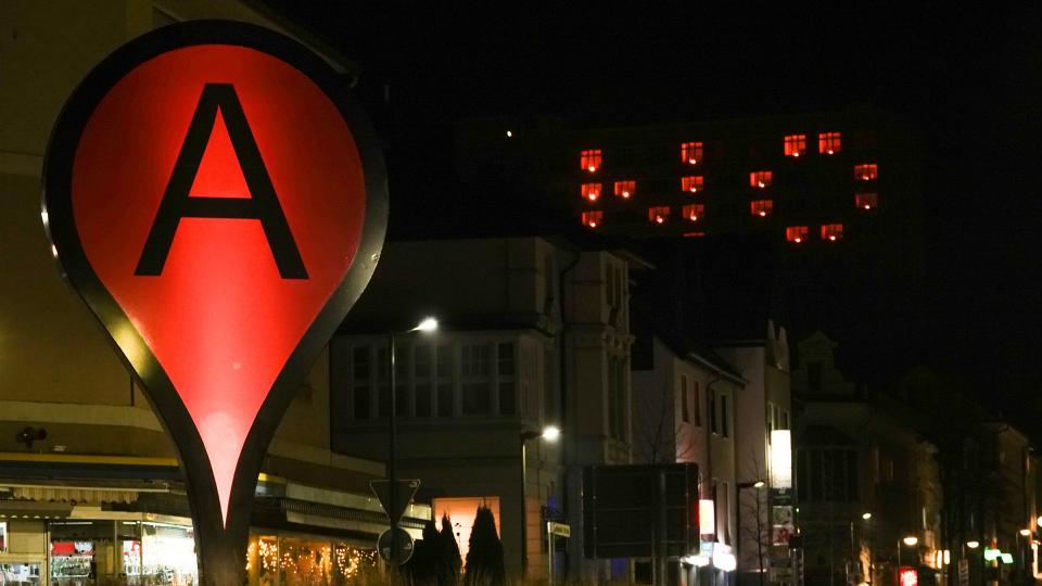 Blick an dem Kunstobjekt "Standort Arnsberg" vorbei auf die Fenster des Gebäudes der Bezirksregierung Arnsberg bei Nacht. Die Fenster sind so beleuchtet, dass die Buchstaben N und O zu lesen sind.