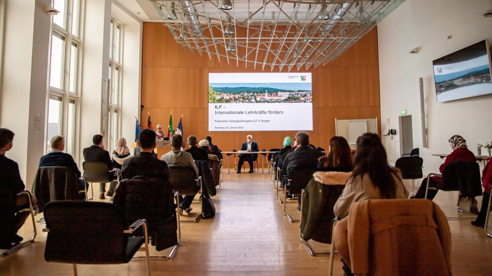 Ein Saal, indem Stühle stehen auf denen Personen sitzen. Sie richten alle Ihren Blick auf eine Person die an einem Tisch sitzt und etwas erzählt. Der Blick ist aus der letzten Reihe.