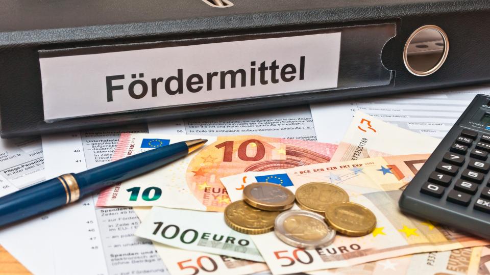 Ein Aktenordner mit der Beschriftung Fördermittel, sowie Geld in Scheinen und Münzen und ein Taschenrechner auf einem Antragsformular.