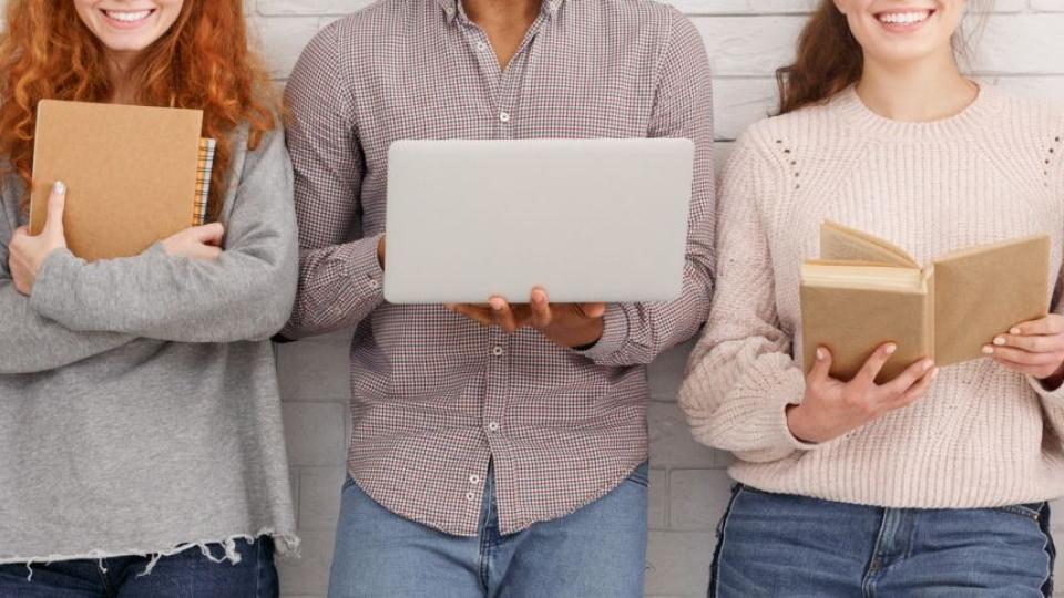 Mehrere junge Erwachsene stehen vor einer weißen Fassade. Sie halten unter anderem einen Laptop, ein Buch und Notizblöcke in den Händen.