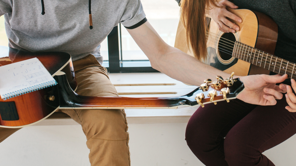 Abgebildet sind zwei Personen, die jeweils eine Gitarre in den Händen halten. Die Person links macht sich zusätzlich Notizen auf einem Blatt Papier. 