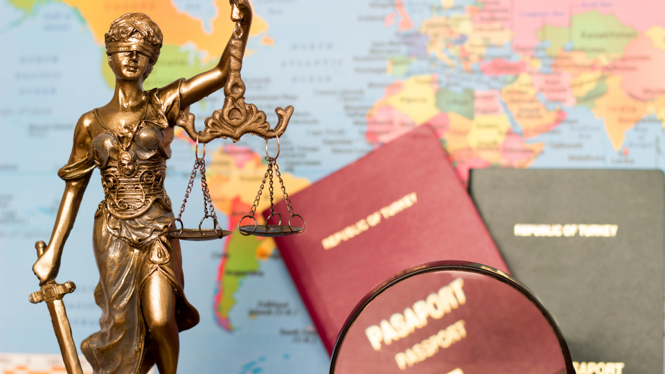 Links abgebildet ist die Figur der Justitia, die Göttin der Gerechtigkeit. Rechts liegen zwei Reisepässe auf einer Weltkarte.