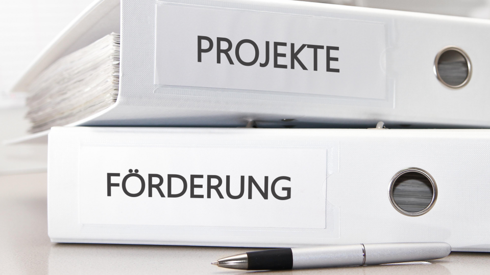 Abgebildet sind ein Kugelschreiber und zwei übereinanderliegende Aktenordner. Der unter Ordner hat die Aufschrift "Förderung" und der obere die Aufschrift "Projekte". 