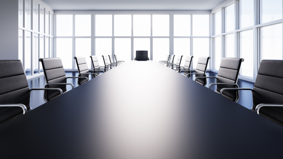 Abgebildet ist ein Konferenzraum mit einem langen dunklen Tisch und jeweils sieben Stühlen rechts und links vom Tisch. Im Mittelpunkt und vor Kopf des Tisches steht ein weiterer Stuhl.