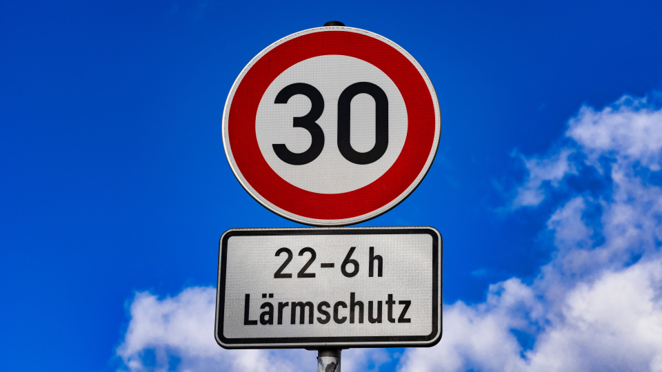 Abgebildet ist ein Tempo-30-Straßenschild. Unter dem Schild ist ein weiteres Schild angebracht, dass darauf hinweist, dass zwischen 22 Uhr und 6 Uhr Lärmschutz beachtet werden muss. 