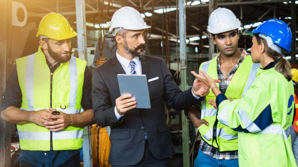 Abgebildet sind vier Personen, die allesamt einen Schutzhelm in einer Industriehalle tragen. Drei der vier Personen tragen zusätzlich eine Warnweste. Der Herr in der Mitte des Bildes trägt einen Anzug und hält ein Tablet in der rechten Hand. Er kommuniziert mit einer Frau neben ihm. 