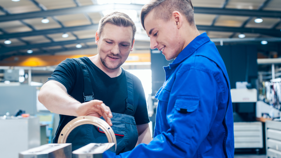 Abgebildet sind zwei Männer in Arbeitskleidung, die sich in einer großen Halle befinden und ein vollständig bearbeitetes Gussteil begutachten. 