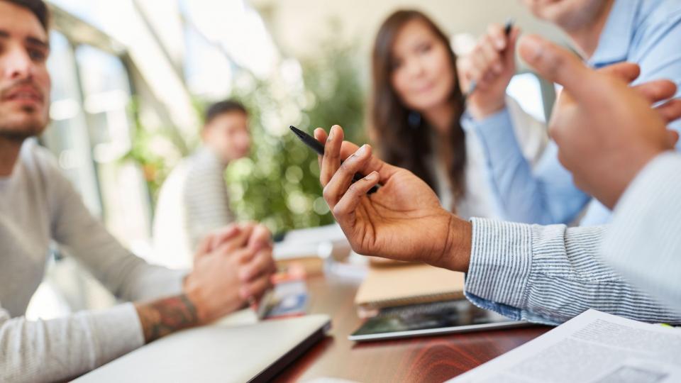 Mehrere Personen sitzen an einem Schreibtisch, auf dem sich ein Laptop, ein Tablet, Schreibblöcke und Schreibutensilien befinden. Die Personen kommunizieren miteinander. 