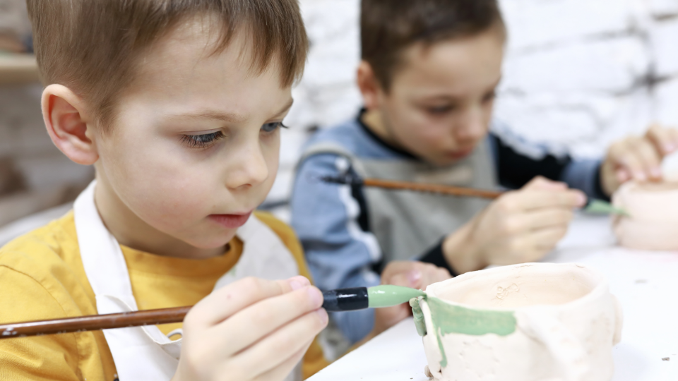 Abgebildet sind zwei Kinder, die Tonfiguren bemalen. 