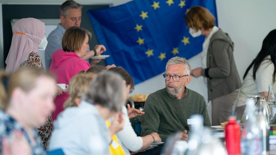 Abgebildet ist ein Seminarraum mit mehreren Personen, die zusammen essen und sich unterhalten. Im Hintergrund hängt die Flagge der Europäischen Union.