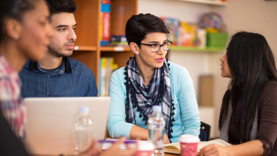 Vier Personen sitzen und unterhalten sich an einem Tisch. Auf dem Tisch liegen Schreibutensilien, Bücher und ein Laptop.