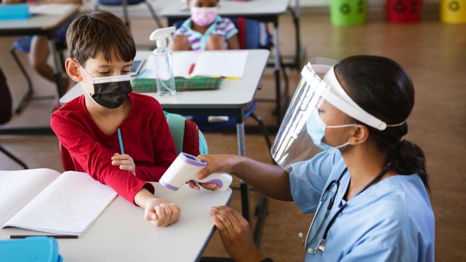 Eine Krankenschwester misst die Temperatur eines Schuljungen in einem Klassenraum. Beide tragen einen Mund- und Nasenschutz.