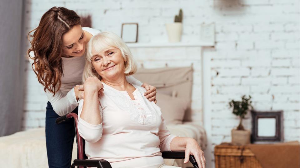 Eine ältere Dame sitzt lächelnd in einem Rollstuhl. Eine jüngere Frau im Hintergrund legt behutsam ihre Hände auf die Schultern der älteren Dame. 