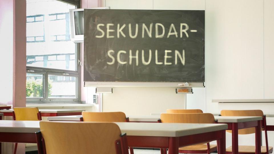 Abgebildet ist ein Klassenraum. Auf der Tafel im Klassenraum steht das Wort "Sekundarschulen".