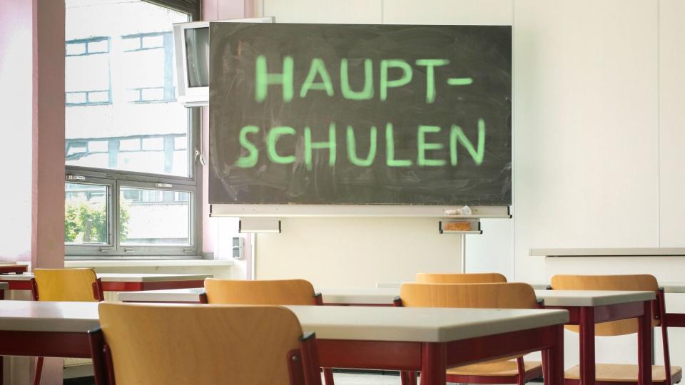 Abgebildet ist ein Klassenraum. Auf der Tafel im Klassenraum steht das Wort "Hauptschulen".