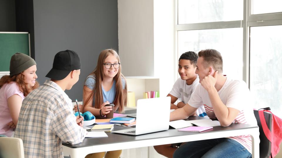 Eine Gruppe Schüler*innen sitzt um einen Tisch. Darauf liegen Bücher und ein aufgeklapptes Notebook.