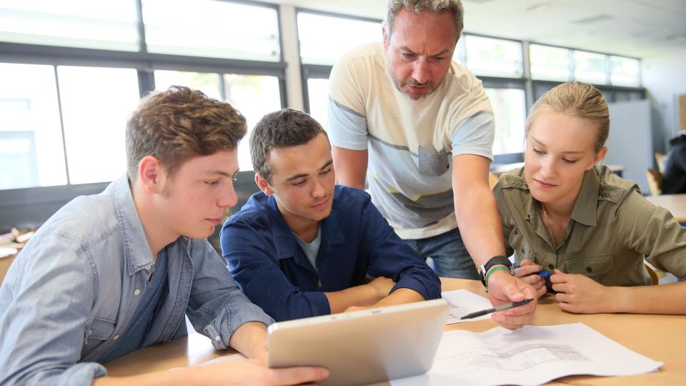 Eine Lehrer und eine Gruppe Schüler*innen mit Tablet und technischer Zeichnung.