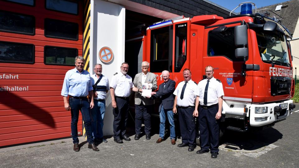 Herr Regierungspräsident Hans-Josef Vogel übergibt einen Förderbescheid an mehrere Vertreter von Stadt und örtlicher Feuerwehr. Alle Personen stehen vor einem Löschfahrzeug, welches zur Hälfte aus der Feuerwehrstation herausgefahren wurde.