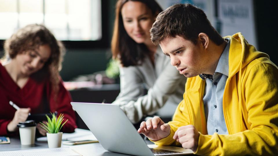 Abgebildet sind drei Personen, die an einem Schreibtisch sitzen. Der junge Mann im Vordergrund hat scheinbar das Downsyndrom und bedient einen Laptop, eine junge Frau im Hintergrund bedient einen Taschenrechner und die dritte Person schaut auf den Laptop. 