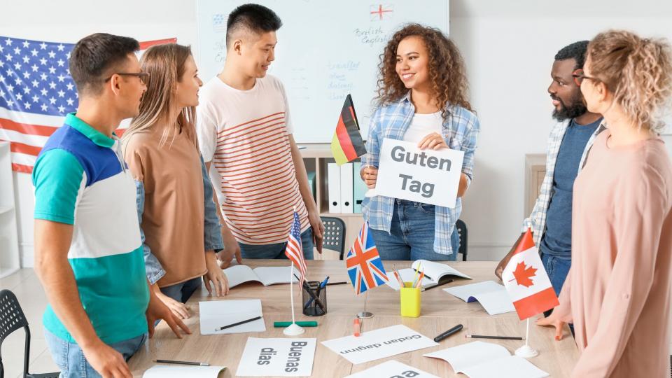 Sechs junge Erwachsene stehen um einen Tisch und schauen auf ein Blatt Papier mit der Aufschrift "Guten Tag", das von einer Frau hochgehalten wird, die gleichzeitig eine Deutschlandflagge in ihrer anderen Hand hält. Auf dem Tisch stehen auch die Flaggen der USA, Großbritannien und Kanada. Die Blätter auf dem Tisch sind in unterschiedlichen Sprachen beschriftet. 